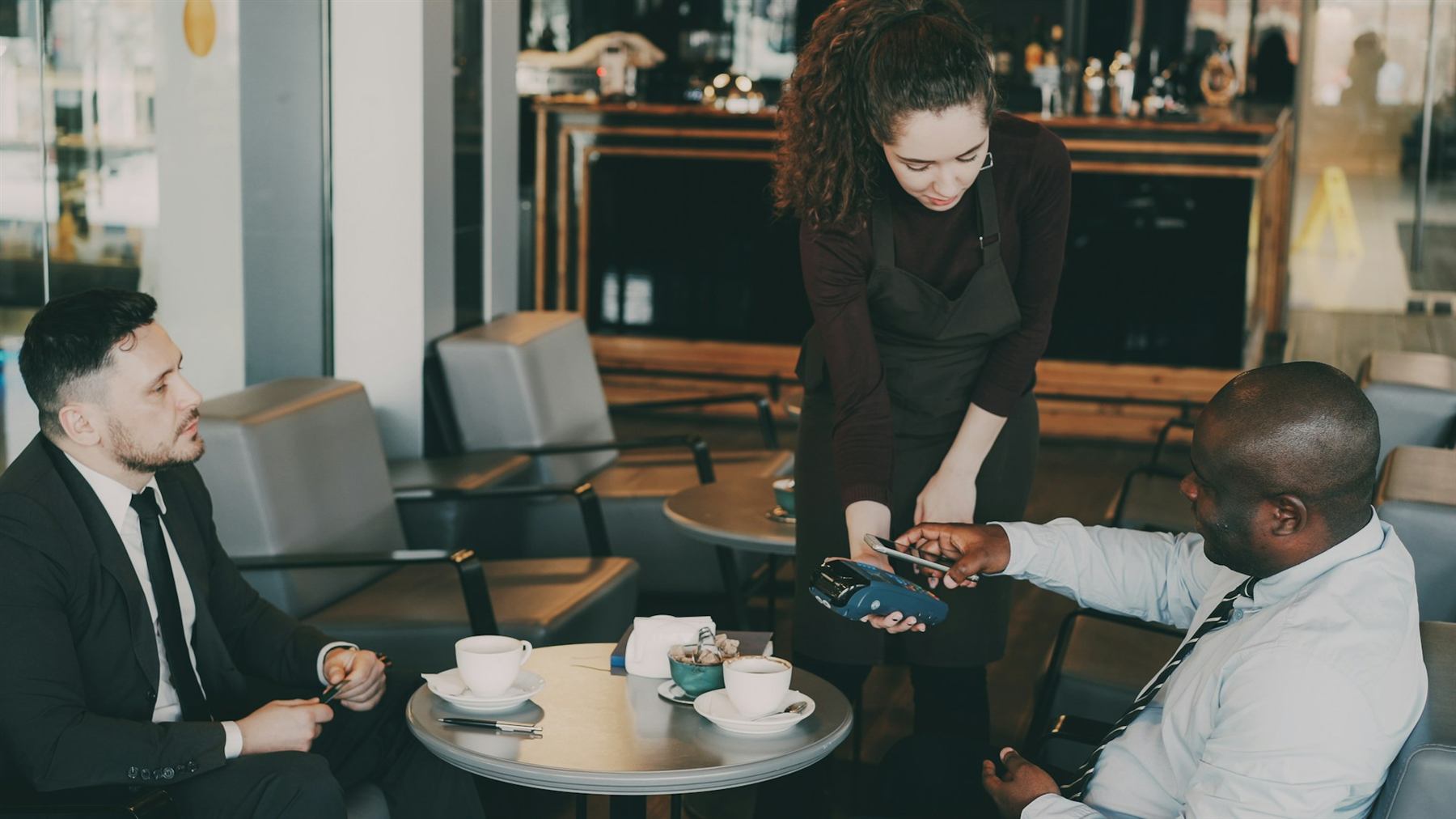Two businessmen paying by phone while a waitress holds a card terminal in a café